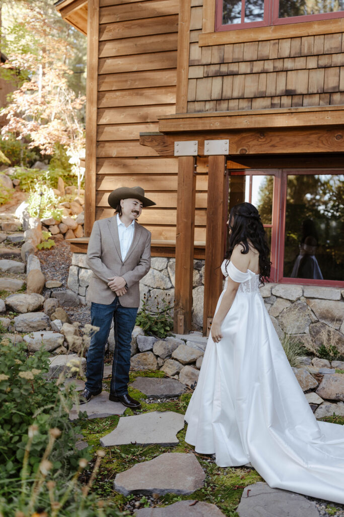 Groom wearing cowboy hat seeing bride during first look on grass with large walking stones in front of wooden building in Lake Tahoe
