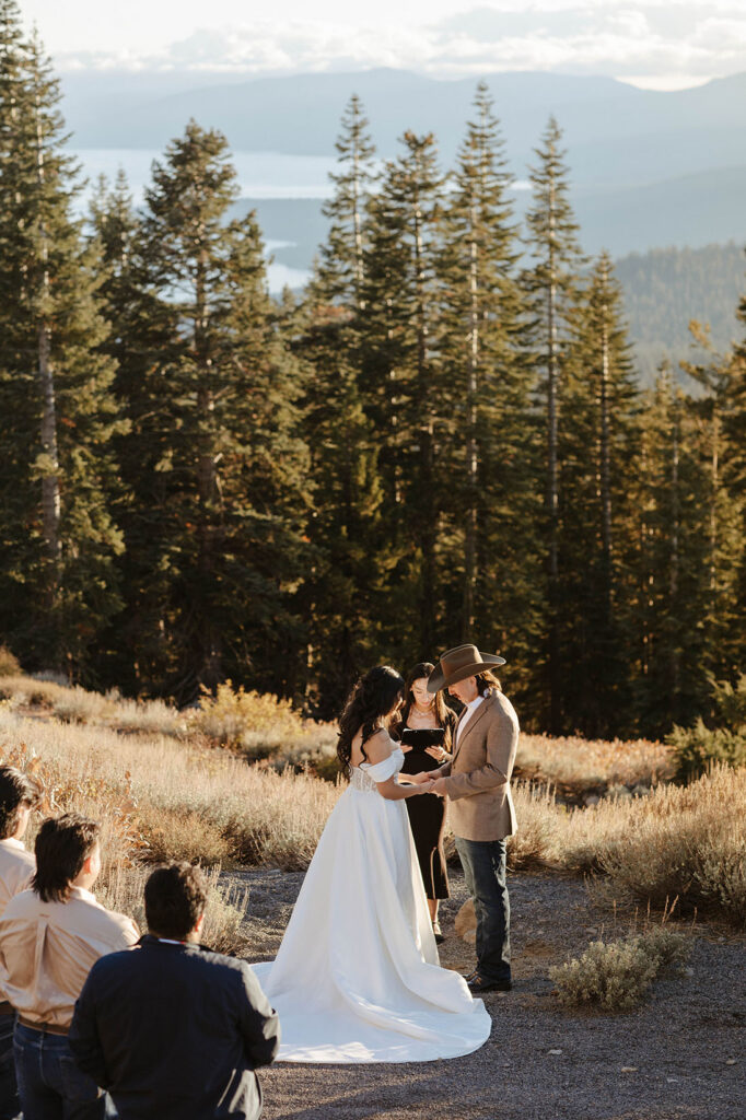 Elopement couple holding hands during ceremony in Lake Tahoe with guests watching and tall pine trees in background