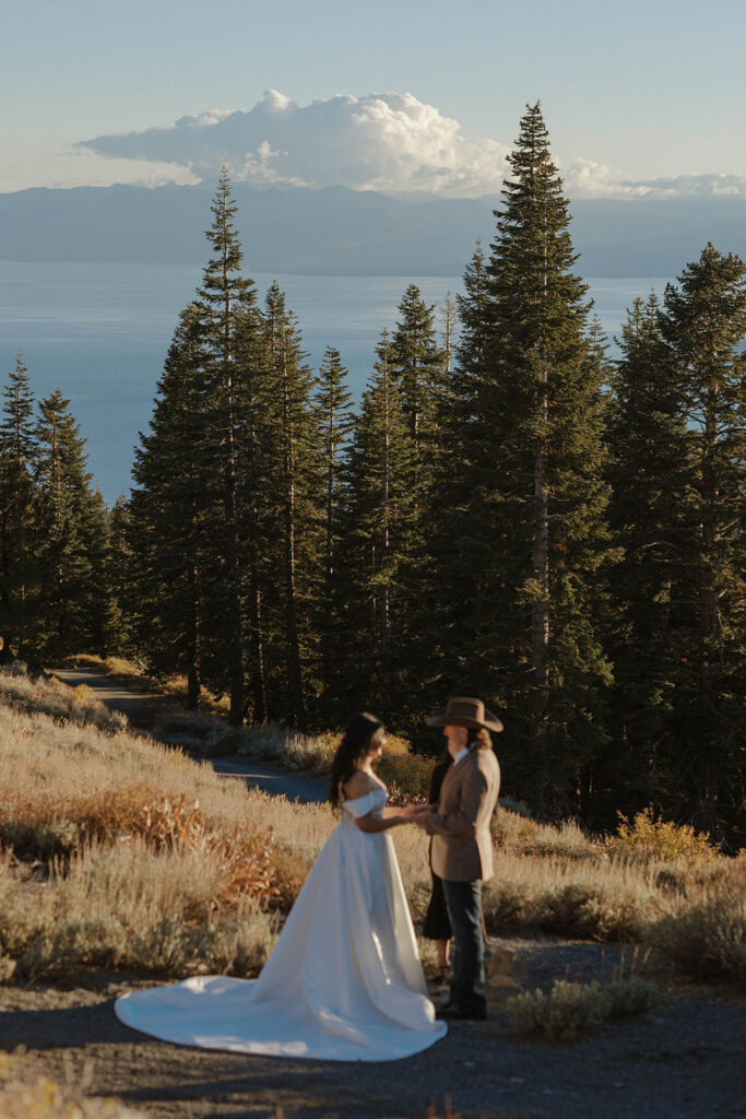 Elopement couple holding hands during ceremony at sunset with tall pine trees and Lake Tahoe in background