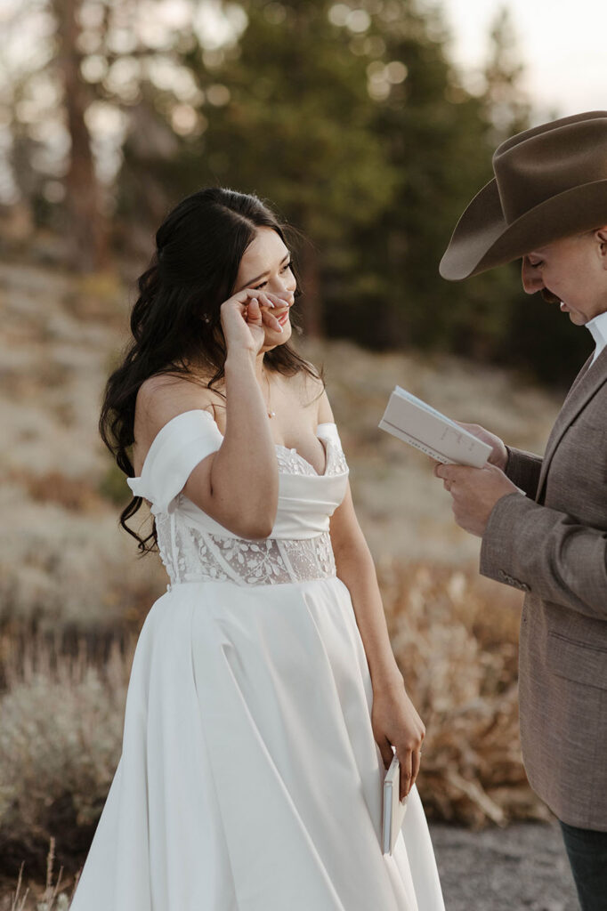 Bride wiping tears from eye while listening to groom reading vows from vow book during elopement ceremony in Lake Tahoe