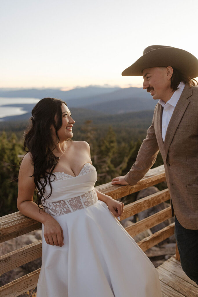 Bride leaning back on wooden railing while smiling at groom in cowboy hat with Lake Tahoe and forest of pine trees in background
