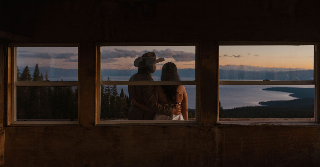 Looking through window at elopement couple with arms around each other and backs to camera looking out at Lake Tahoe