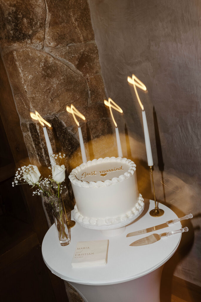 White wedding cake with gold accents and cutlery next to white florals and candles 