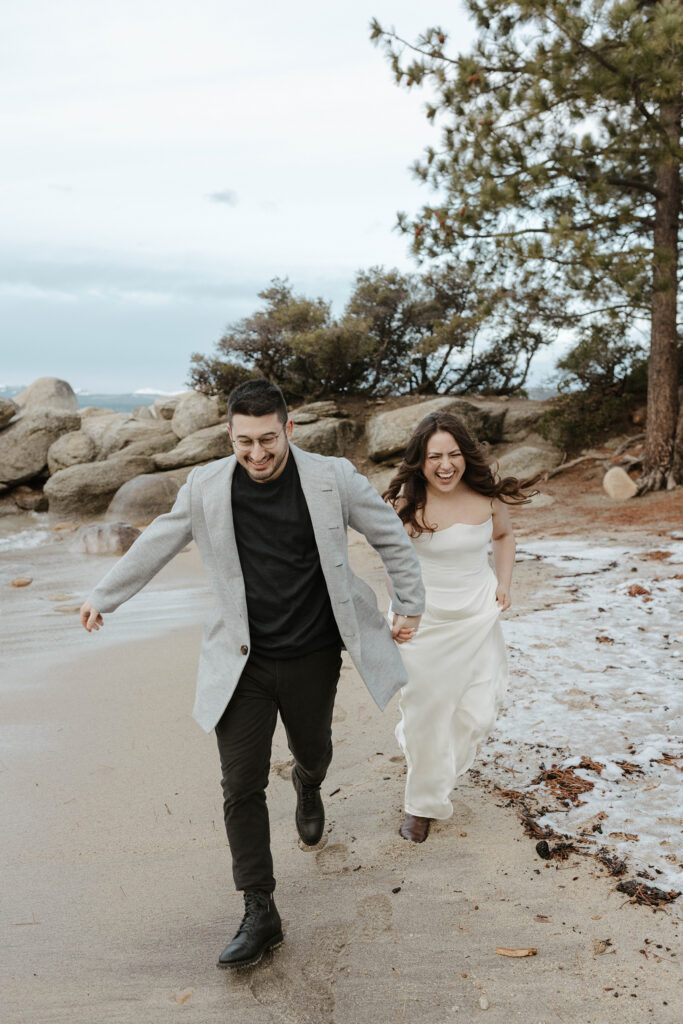 Engagement couple holding hands while running across Lake Tahoe beach in winter while laughing with large rocks in background