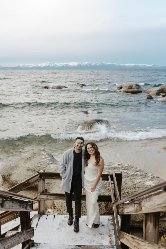 Engagement couple standing together on wooden staircase on snow and smiling at camera with Lake Tahoe and beach behind them
