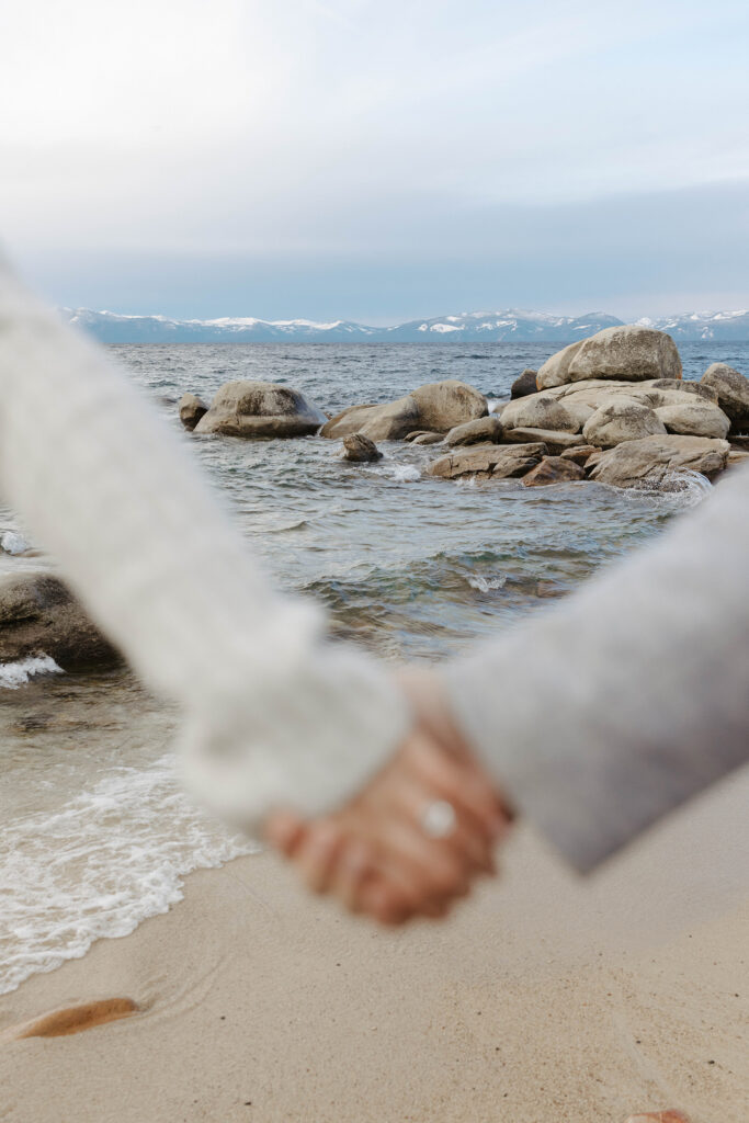 Close up of engagement couple holding hands with engagement ring on woman's finger with Lake Tahoe and large rocks in background