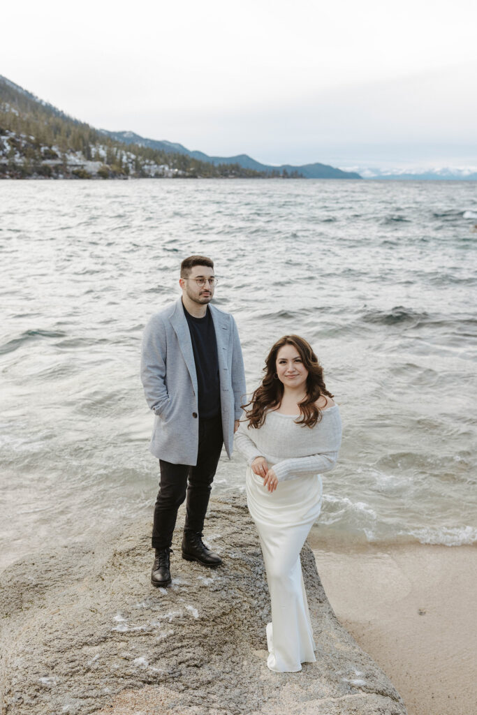 Engagement couple standing on boulder next to each other on a beach during winter with Lake Tahoe in background