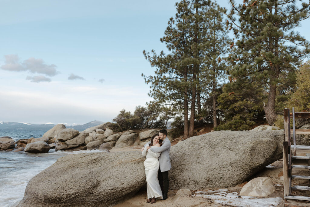 Man hugging fiancé from behind while couple stands in front of large boulders next to Lake Tahoe with pine trees in background