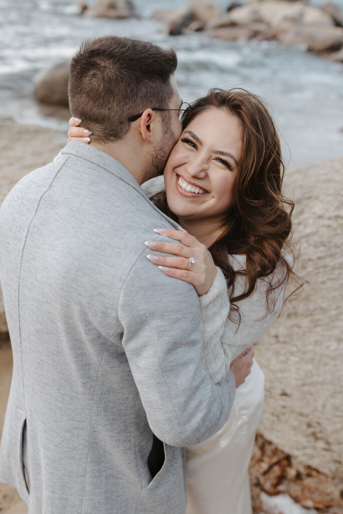 Engagement couple hugging each other while woman smiles at camera while on a beach in Lake Tahoe