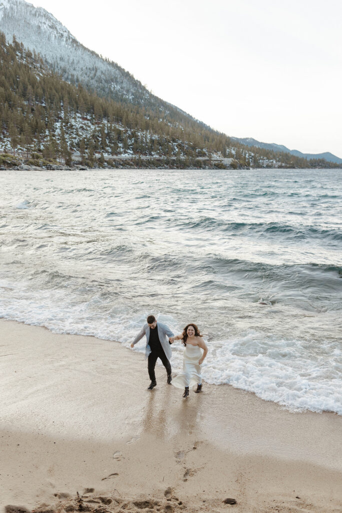 Engagement couple holding hands while running up sandy beach with Lake Tahoe and snowy pine trees in background