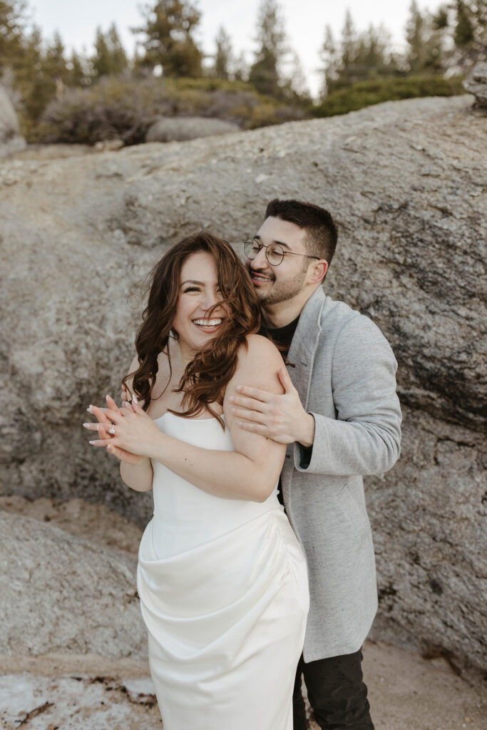 Man hugging fiancé from behind while she rubs her hands together to stay warm and smiles while in Lake Tahoe with large rocks behind them