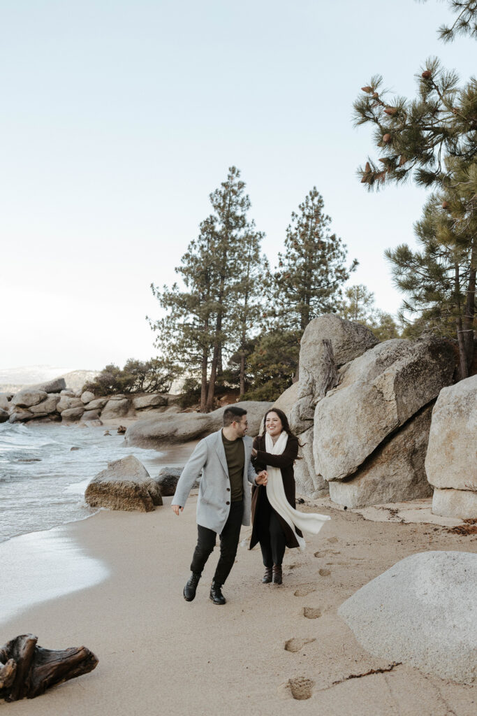 Engagement couple holding hands while running along Lake Tahoe beach together and smiling at each other with large rocks behind them