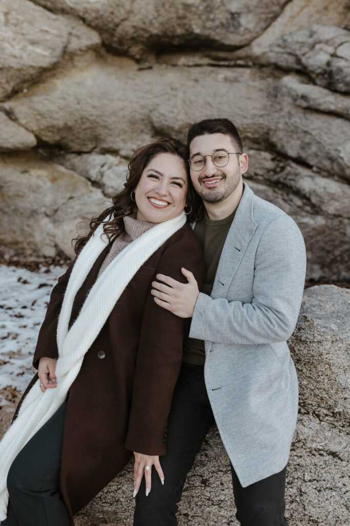 Engagement couple sitting on boulder together and smiling at camera with large rocks and snow in background in Lake Tahoe