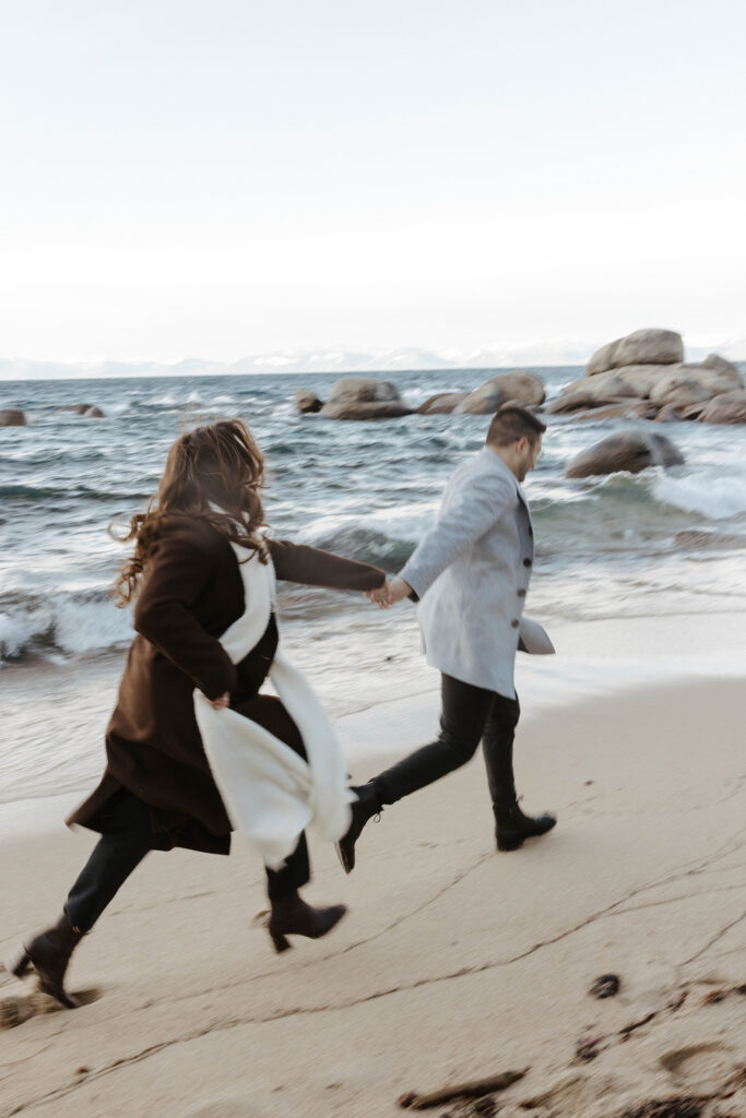 Engagement couple holding hands and running along sandy beach together away from camera with Lake Tahoe next to them