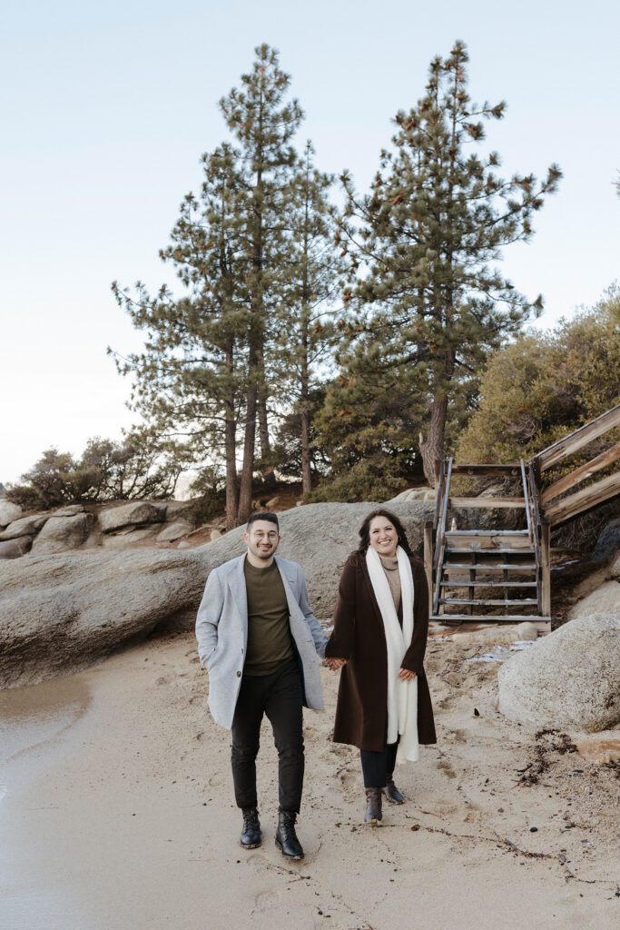 Engagement couple smiling at camera while standing on sandy beach together and holding hands in Lake Tahoe with large rocks and wooden stairs in background