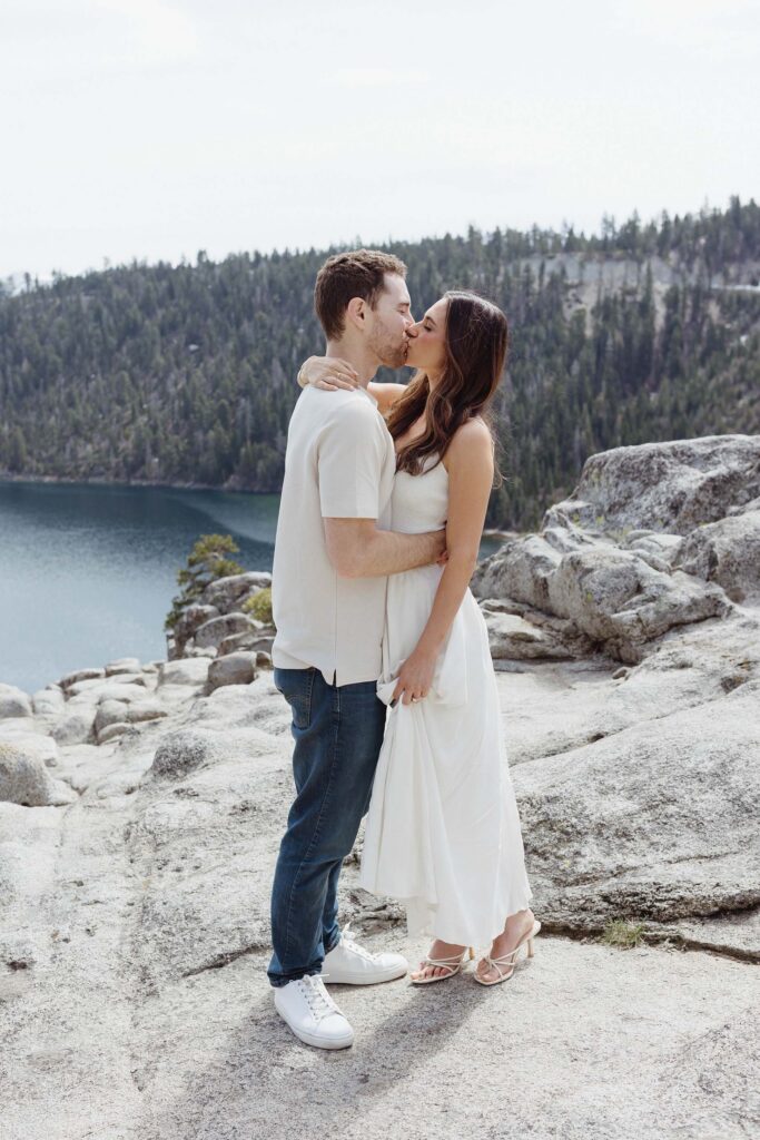 Engagement couple holding each other and kissing while standing on rocky cliff with Lake Tahoe and lots of pine trees in background in Emerald Bay
