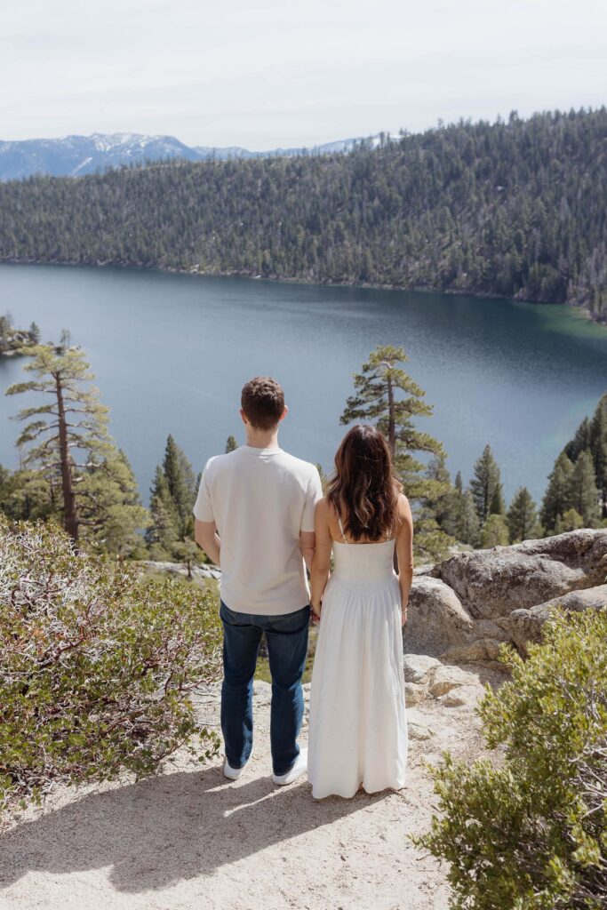 Engagement couple holding hands while looking out over Lake Tahoe and forest of pine trees at Emerald Bay
