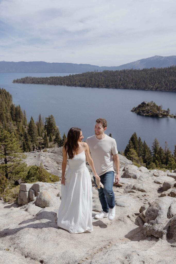 Engagement couple walking up rocky path and smiling at each other with Lake Tahoe in background at Emerald Bay
