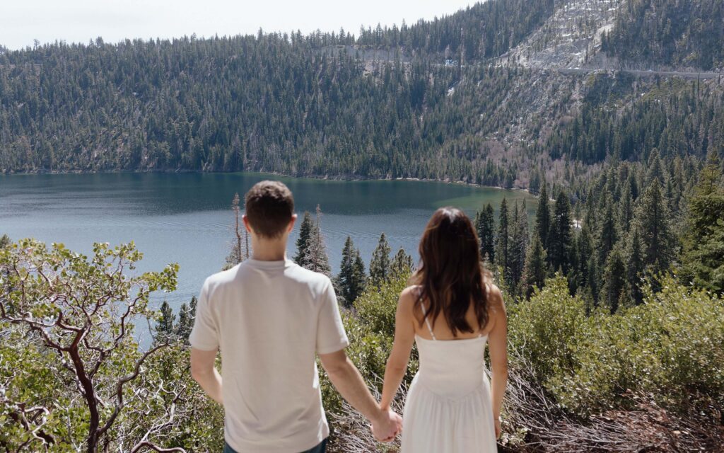 Engagement couple holding hands while looking at Lake Tahoe together next to lots of greenery at Emerald Bay