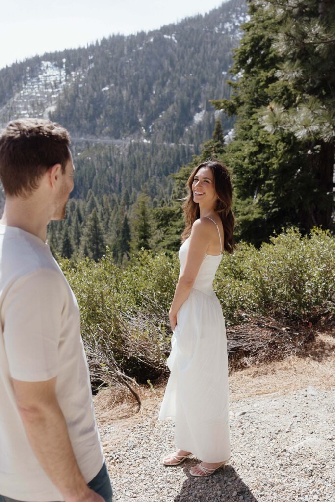 Woman looking over at fiancé and smiling with lots of pine trees in background at Emerald Bay