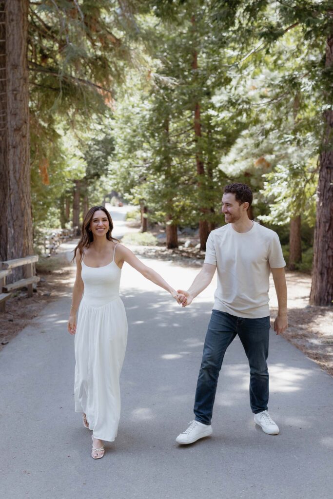 Engagement couple smiling while holding hands and walking along path together surrounded by tall pine trees at Emerald Bay