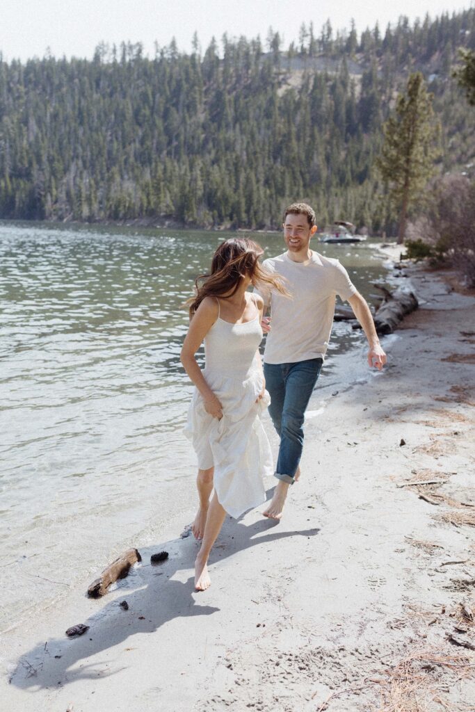 Engagement couple running along sandy beach together bare foot while woman looks back at fiancé in Emerald Bay