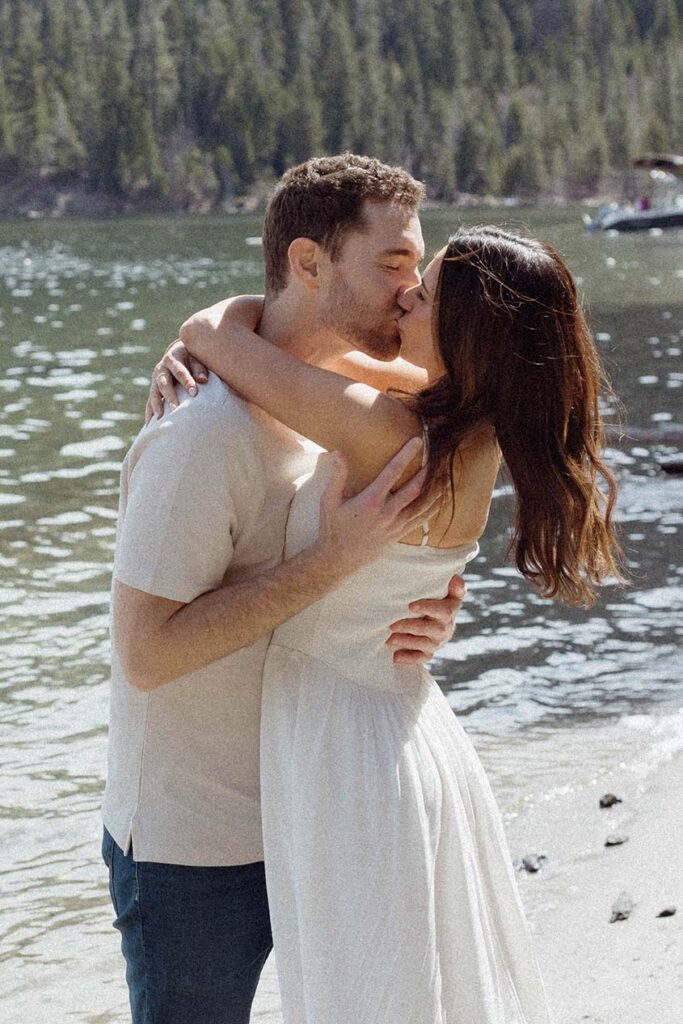 Engagement couple holding each other and kissing while on sandy beach with Lake Tahoe in background at Emerald Bay