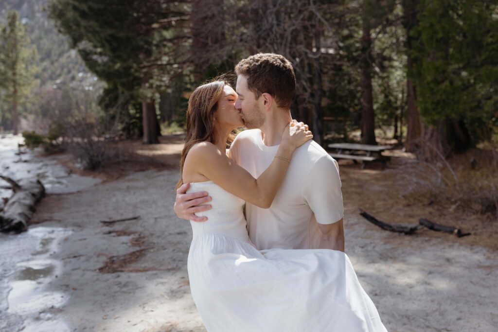 Man holding fiancé while couple is kissing with tall pine trees in background at Emerald Bay