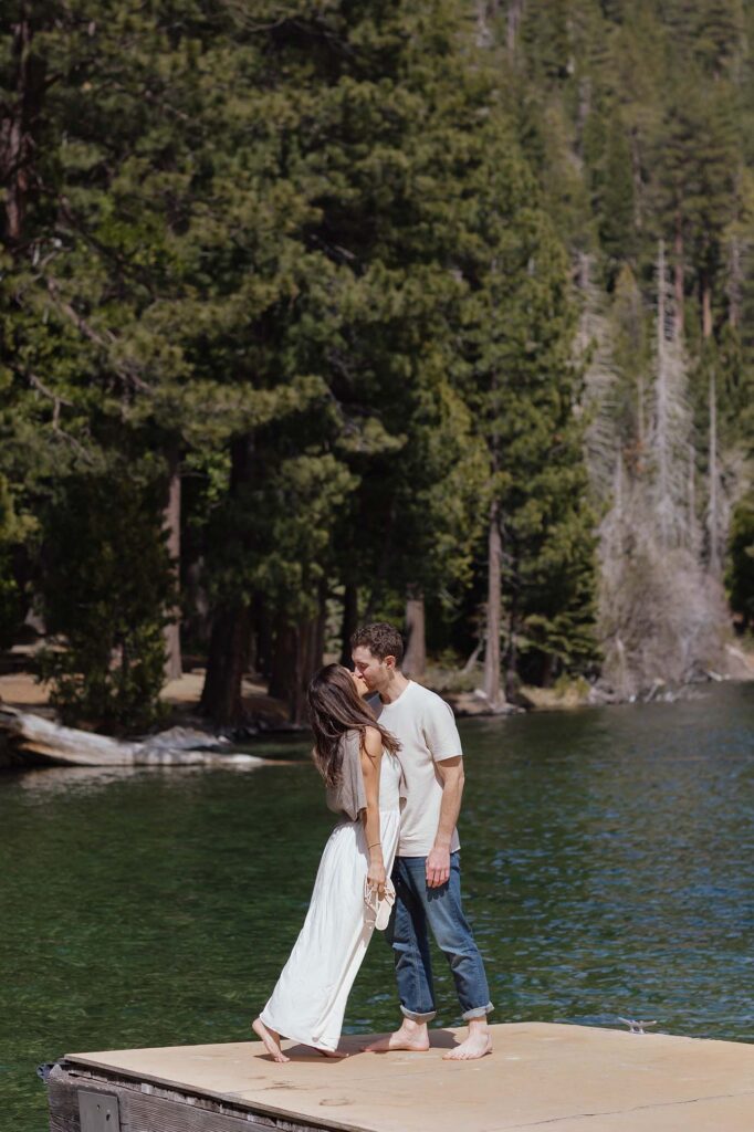 Engagement couple standing on end of pier together and kissing with tall pine trees in background in Emerald Bay