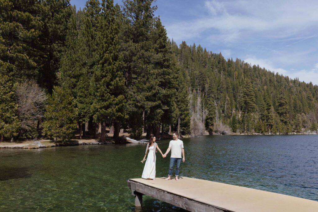Engagement couple holding hands while standing on end of pier together in Emerald Bay with tall pine trees in background