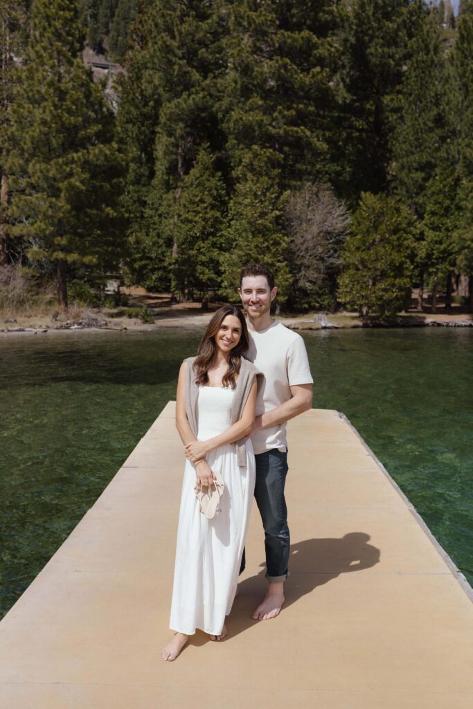 Engagement couple standing together on a pier in Emerald Bay while smiling at camera with pine trees in background