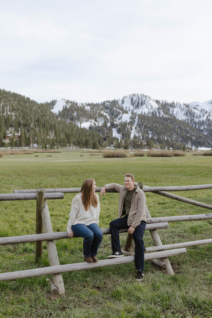 Engagement couple sitting on wooden fence together in open grass field with snowy mountains in background at Palisades 