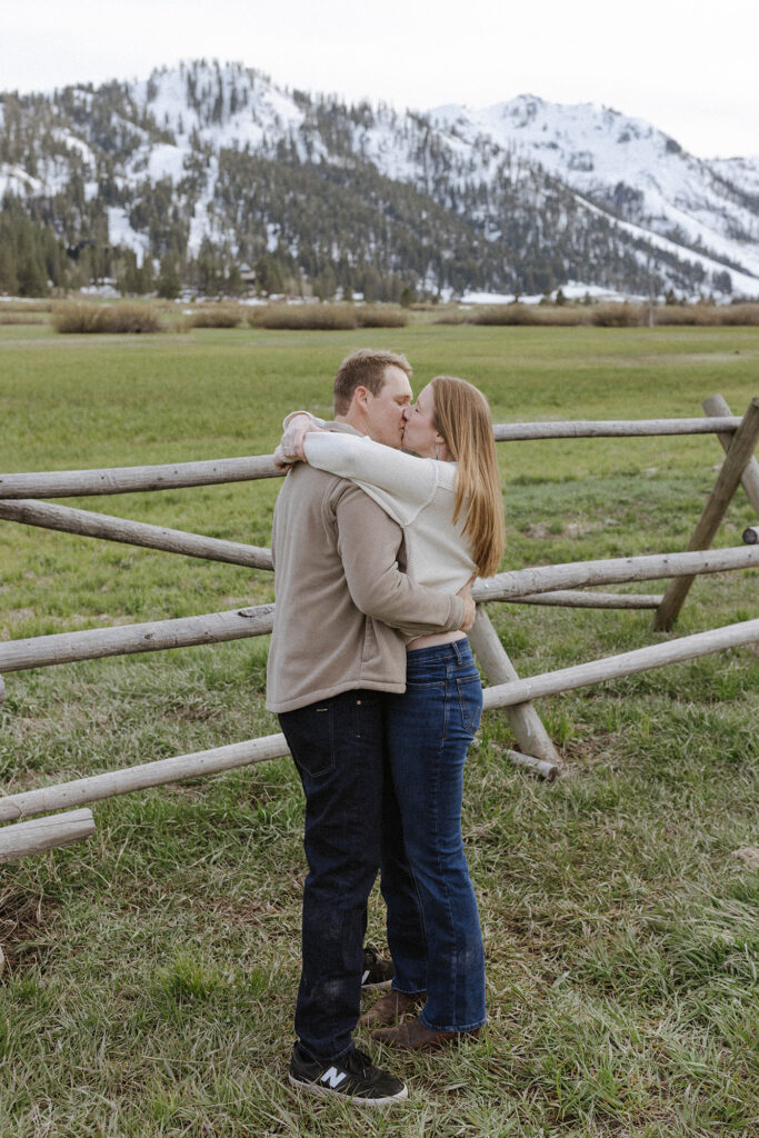 Engagement couple hugging and kissing in front of wooden fence in open field with snowy mountains in background at Palisades