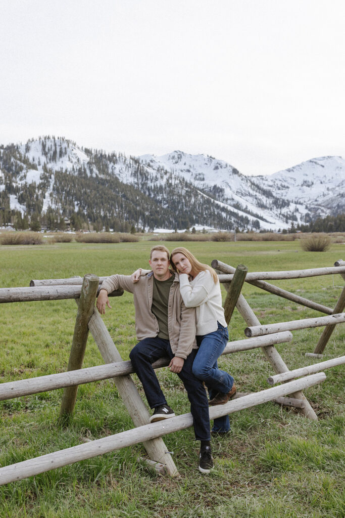 Engagement couple sitting on wooden fence together while woman leans on fiancé's shoulder with snowy mountains in background at Palisades