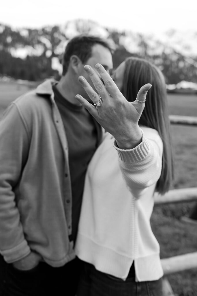 Woman showing off engagement ring to camera while kissing fiancé with snowy mountains in background at Palisades