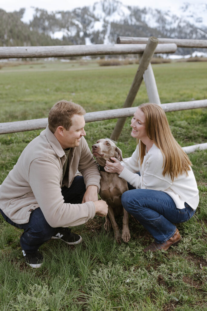 Engagement couple crouching down and holding their dog while smiling in front of wooden fence with snowy mountains in background at Palisades