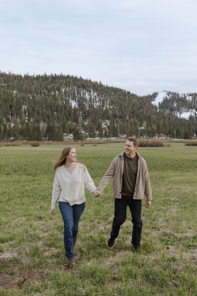 Engagement couple holding hands while walking across open field and smiling at each other with snowy mountains in background at Palisades