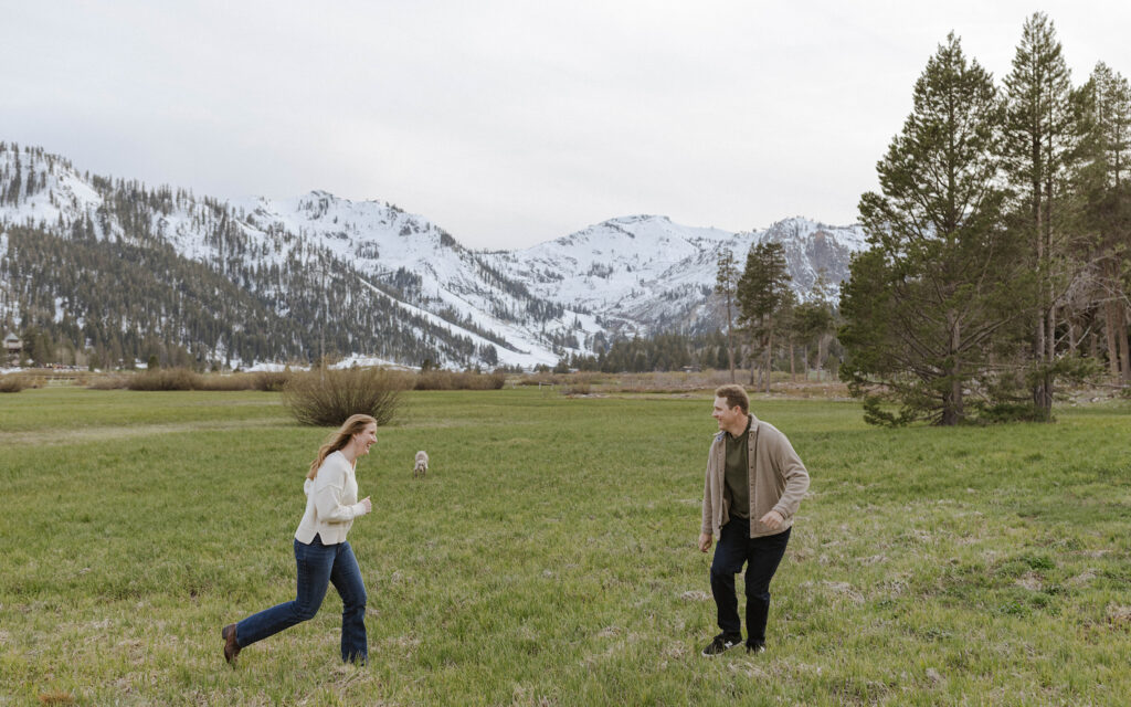 Woman running towards fiancé across open field with their dog and snowy mountains in background at Palisades