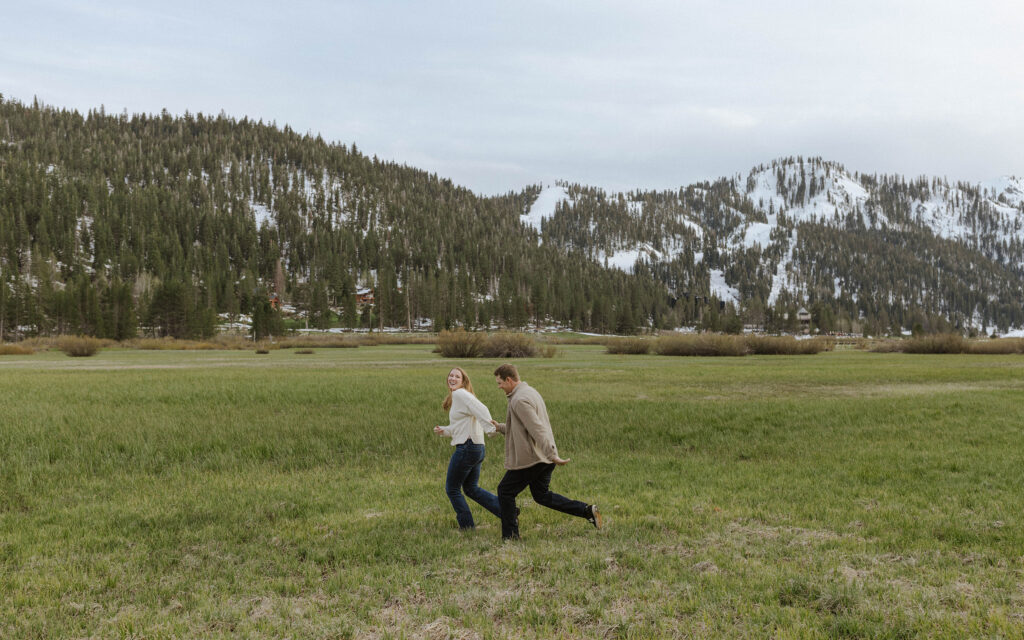 Engagement couple holding hands and running across field together while woman looks back over shoulder and laughs with snowy mountains in background at Palisades