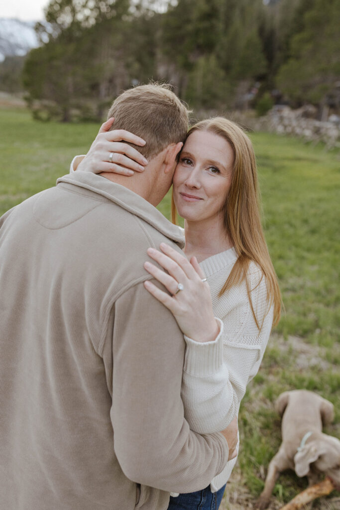 Woman holding her fiancé and hugging him while smiling at camera with dog and open field in background at Palisades