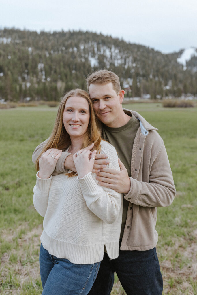Engagement couple holding each other while smiling at camera with snowy mountains in background at Palisades