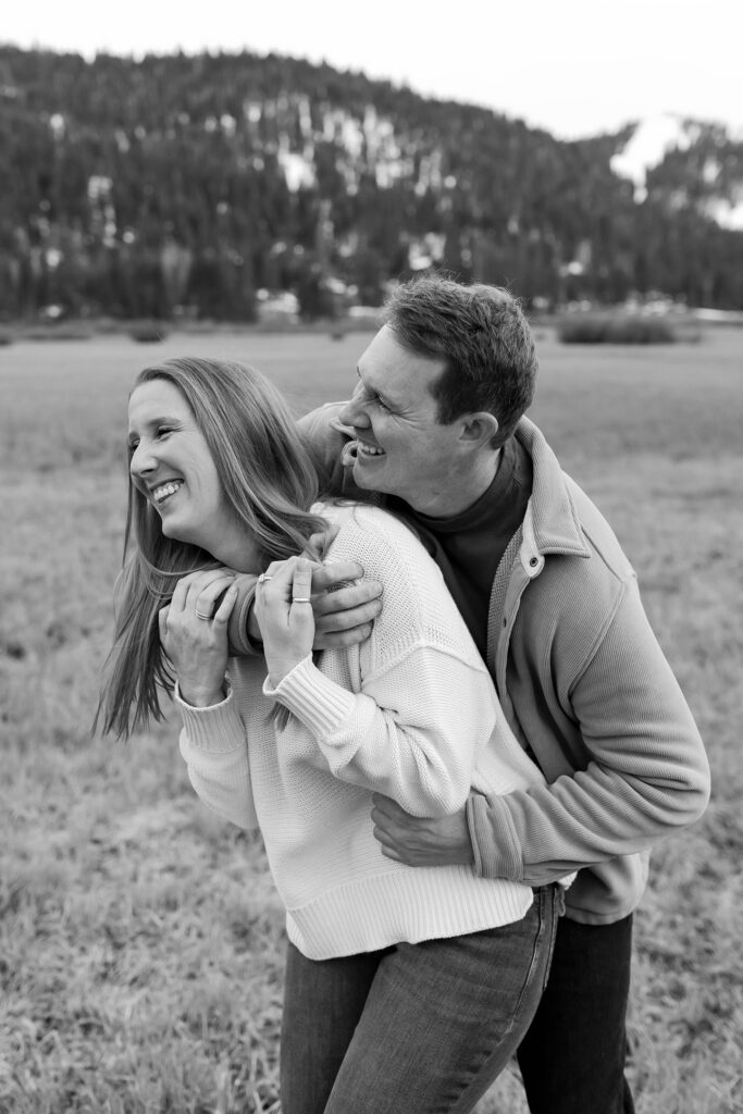 Man hugging fiancé from behind while both are laughing with snowy mountains in background at Palisades