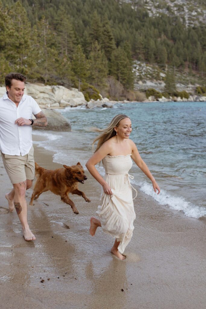 Engagement couple running along sandy beach barefoot next to Lake Tahoe with their dog while woman holds dress and pine trees in background