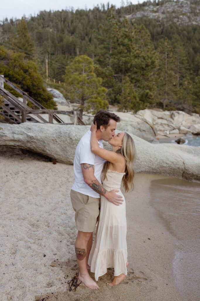 Engagement couple holding each other and kissing while on a sandy beach next to Lake Tahoe with large rocks and pine trees in background