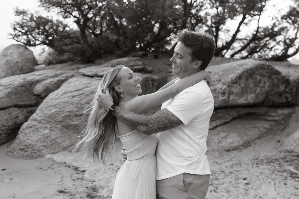 Engagement couple holding and smiling at each other while on a beach in Lake Tahoe with tall rocks behind them