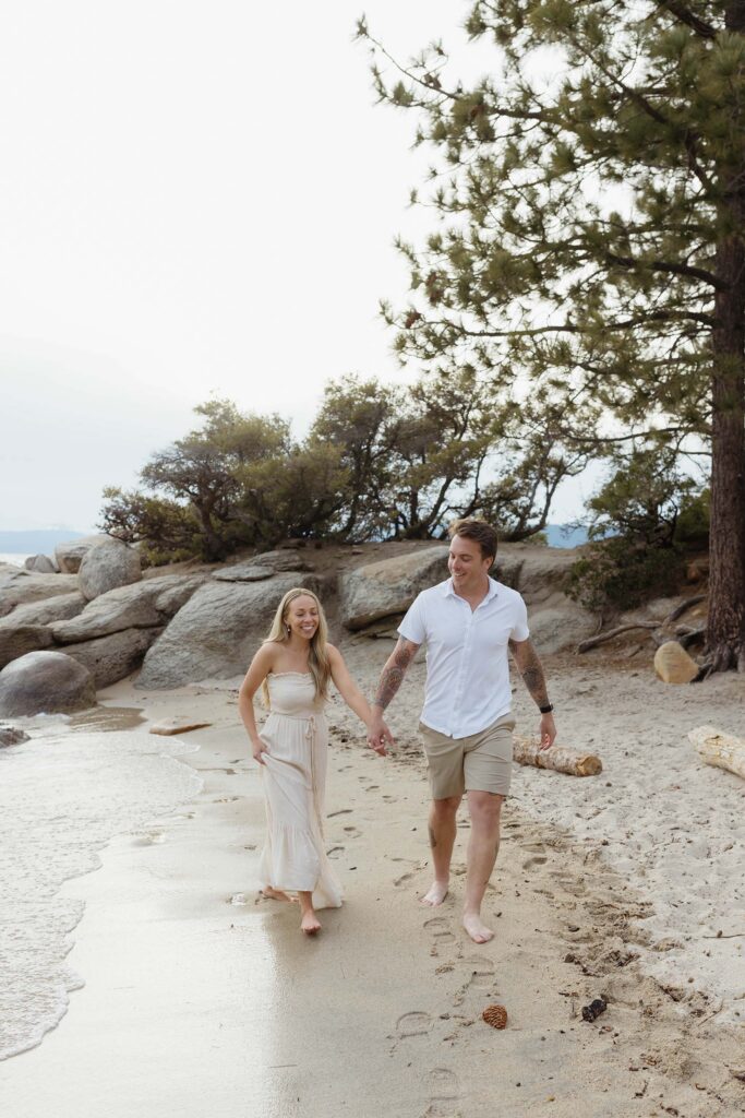 Engagement couple holding hands while smiling and walking along sandy beach together next to Lake Tahoe