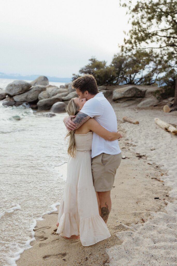 Engagement couple hugging each other while standing on sandy beach next to waves in Lake Tahoe with large rocks in background