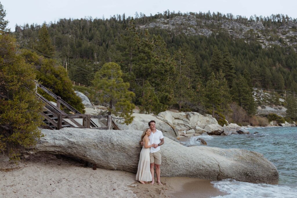 Engagement couple holding each other while leaning on a large rock on a beach in Lake Tahoe with lots of pine trees in background