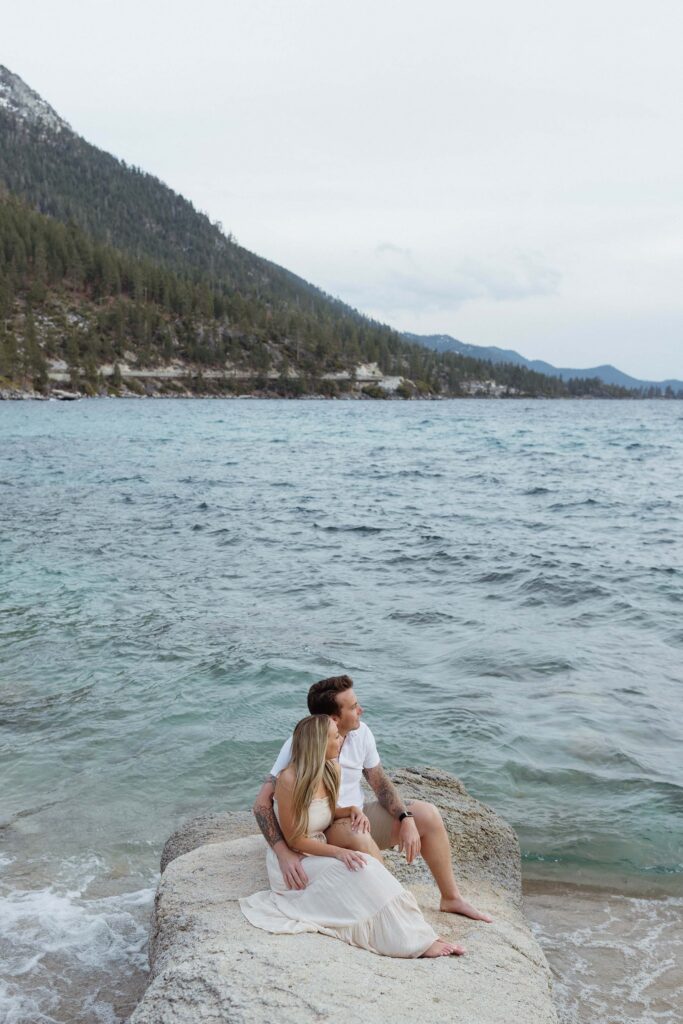 Engagement couple sitting on large rock together while looking out at Lake Tahoe