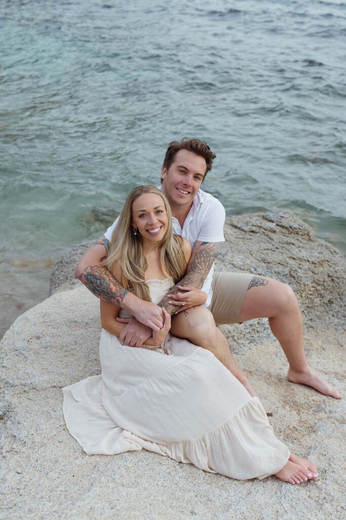 Engagement couple holding each other while sitting on a large rock together and smiling at camera with Lake Tahoe in background